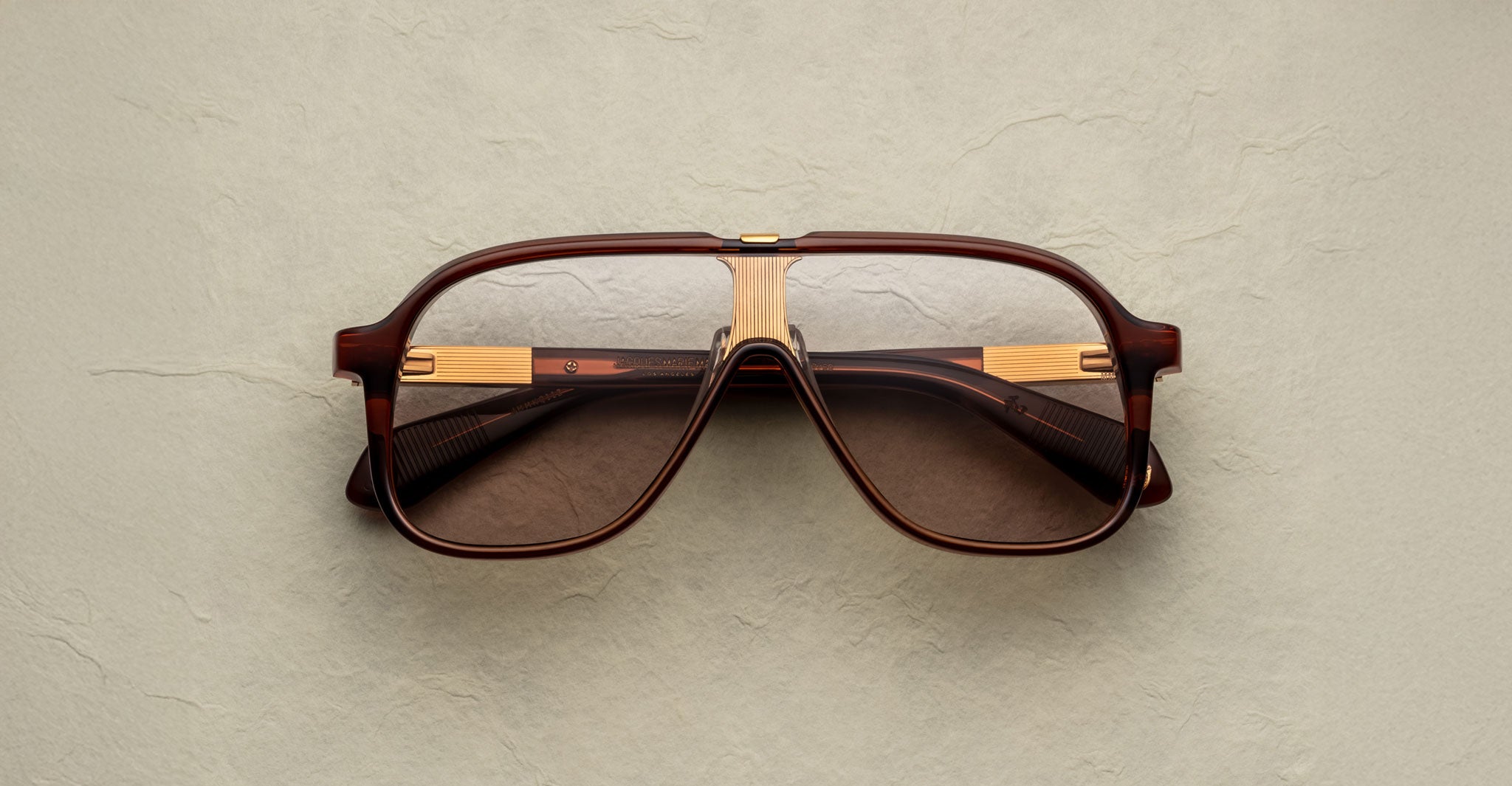 The Jacques Marie Mage Kurokawa vintage-style brown and gold aviator eyeglasses with large lenses are displayed on a textured beige surface, viewed from the front.