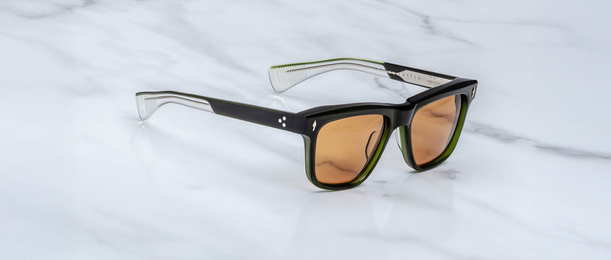 The Jacques Marie Mage Lankaster sunglasses, featuring thick black and transparent frames with brown-tinted lenses, rest on a white marble surface.
