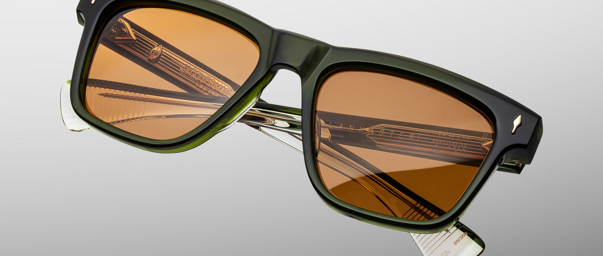 The Jacques Marie Mage Lankaster rectangular sunglasses feature dark green frames, brown-tinted lenses, and metallic inner details on the folded temples, displayed against a gray gradient background.