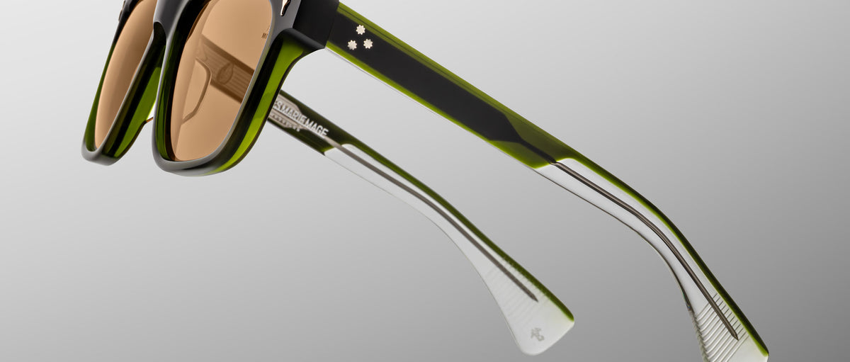 The Jacques Marie Mage Lankaster sunglasses feature thick, square black and green frames, brown-tinted lenses, transparent arm tips, and the brand name visible on the inner side of the arm.