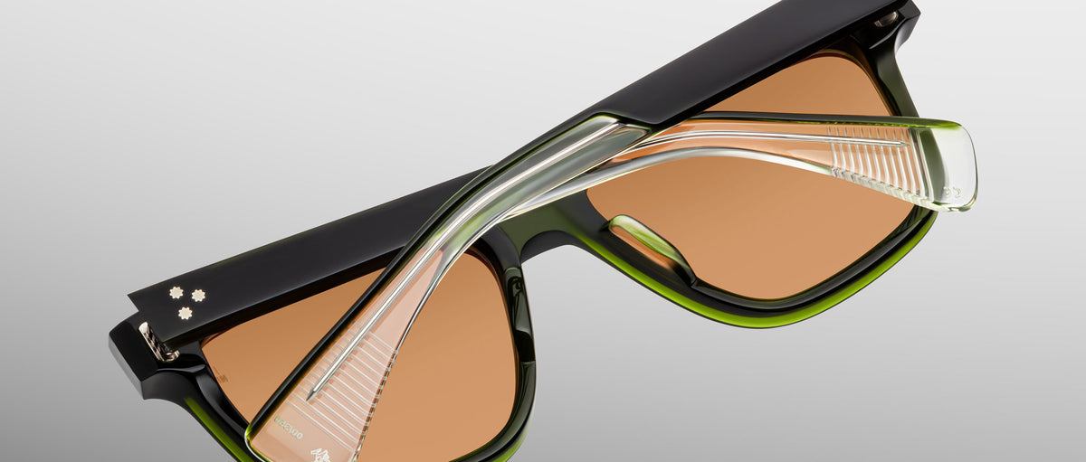 A close-up of Jacques Marie Mage Lankaster sunglasses, featuring thick black and green frames, brown-tinted lenses, and transparent temples set against a neutral gradient background.