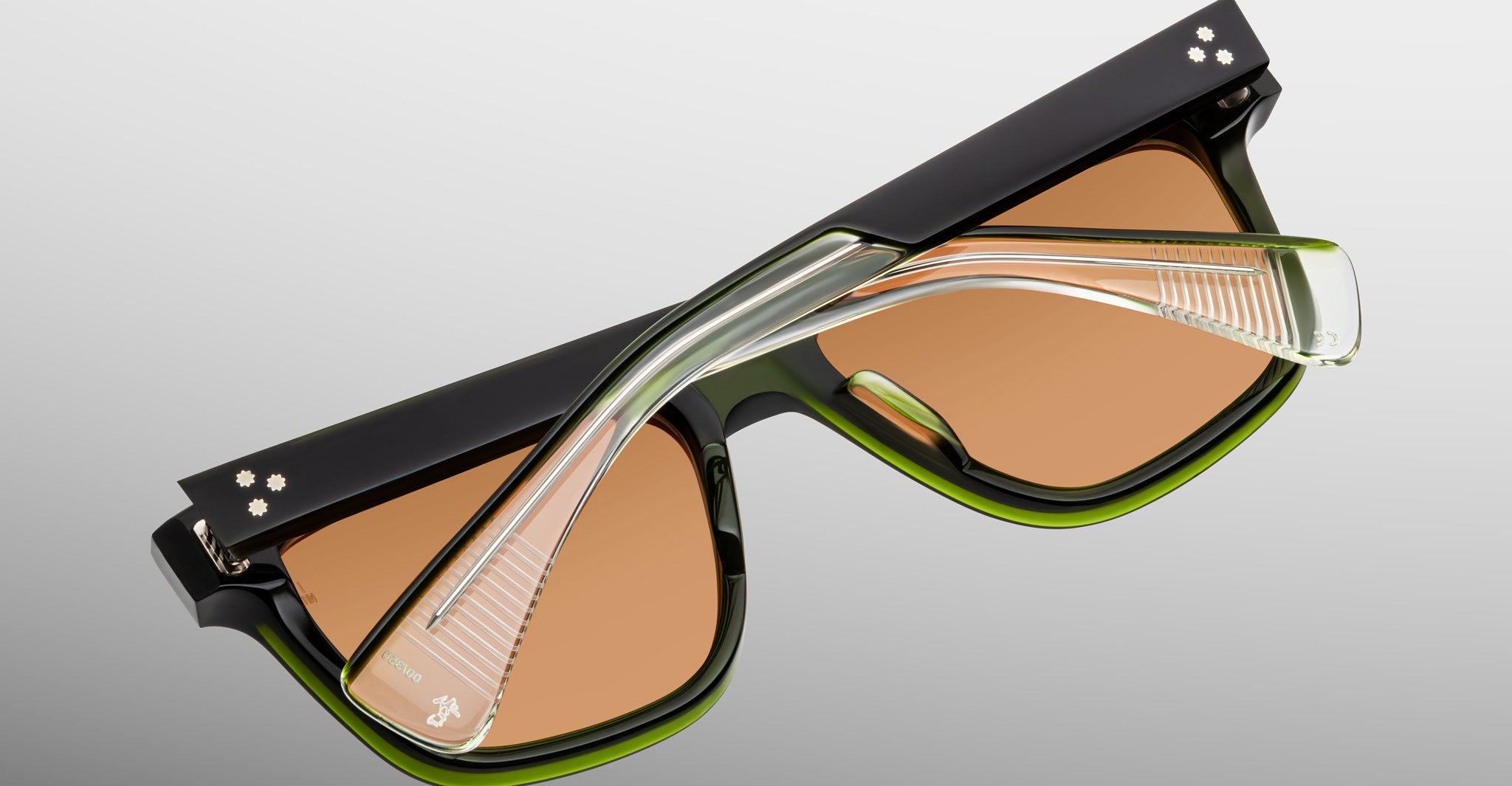 A close-up of Jacques Marie Mage Lankaster sunglasses, featuring thick black and green frames, brown-tinted lenses, and transparent temples set against a neutral gradient background.