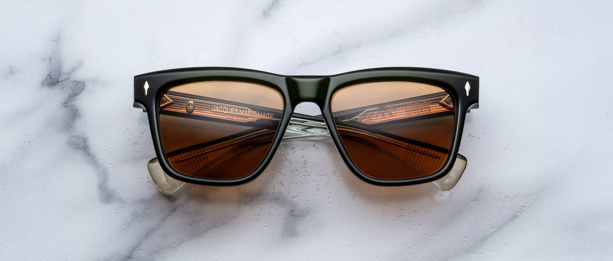 A pair of Jacques Marie Mage Lankaster black rectangular sunglasses with brown lenses lies on a white marble surface, the brand text visible inside the arms and a striped pattern reflected in the lenses.