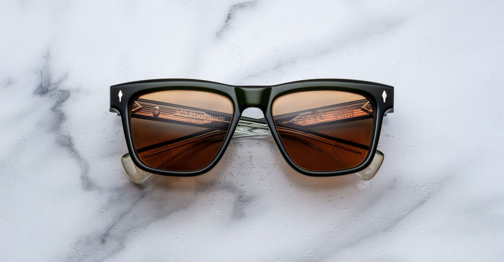 A pair of Jacques Marie Mage Lankaster black rectangular sunglasses with brown lenses lies on a white marble surface, the brand text visible inside the arms and a striped pattern reflected in the lenses.