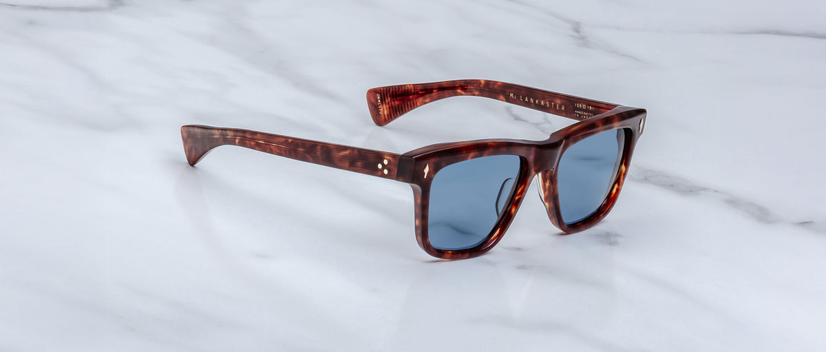 A pair of Jacques Marie Mage Lankaster sunglasses with square dark tortoiseshell frames and blue-tinted lenses rests on a white marble surface.