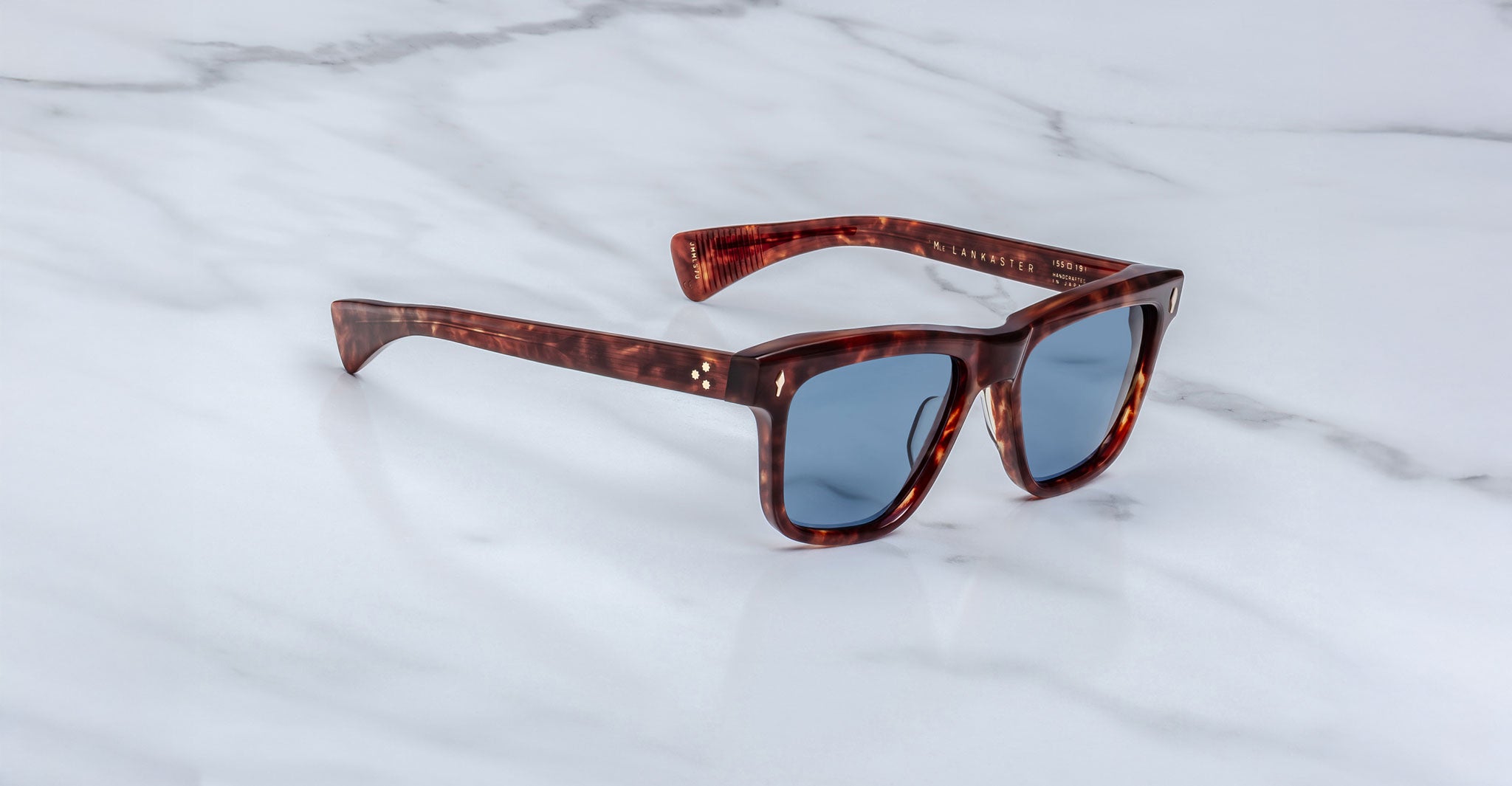 A pair of Jacques Marie Mage Lankaster sunglasses with square dark tortoiseshell frames and blue-tinted lenses rests on a white marble surface.