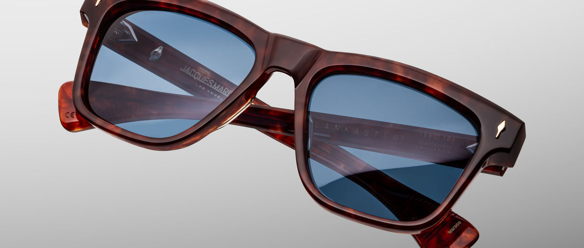 The Jacques Marie Mage Lankaster is a pair of square-framed tortoiseshell sunglasses with blue-tinted lenses, displayed against a plain gray background. The arms feature inscribed branding text.