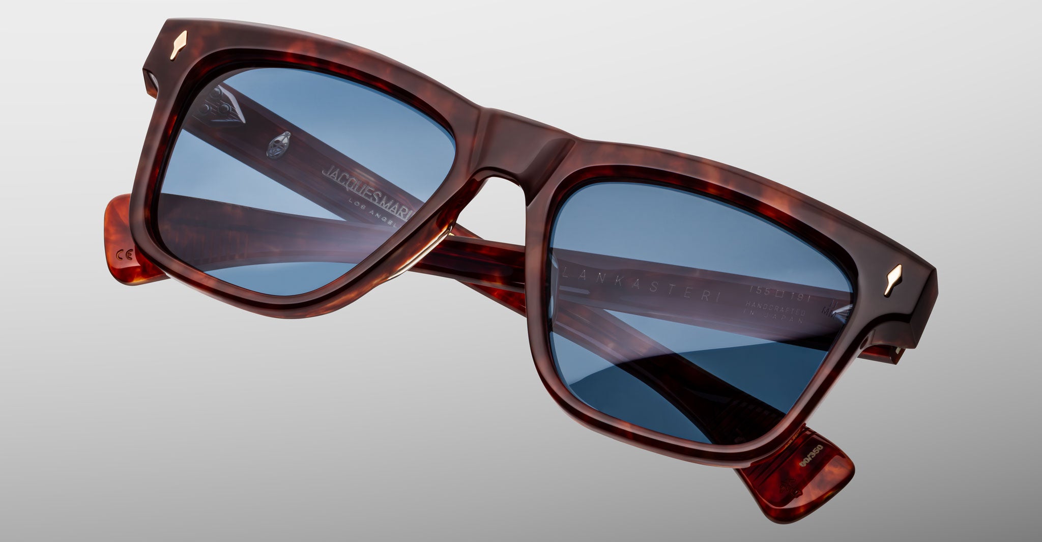 The Jacques Marie Mage Lankaster is a pair of square-framed tortoiseshell sunglasses with blue-tinted lenses, displayed against a plain gray background. The arms feature inscribed branding text.