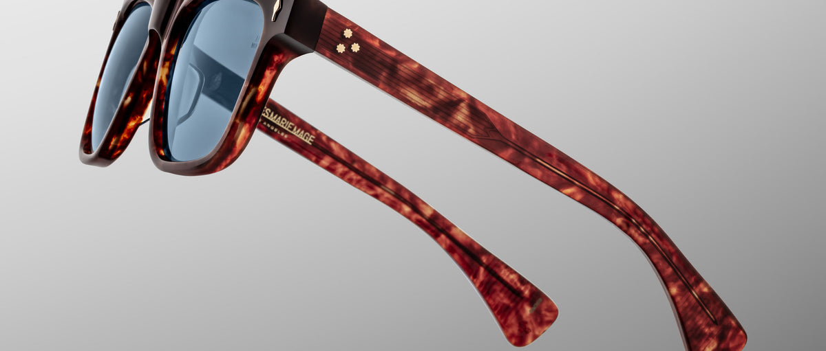 The Jacques Marie Mage Lankaster sunglasses feature a tortoiseshell pattern, blue lenses, thick frames, and gold accents on the arms, shown from a side angle against a plain light gray background.