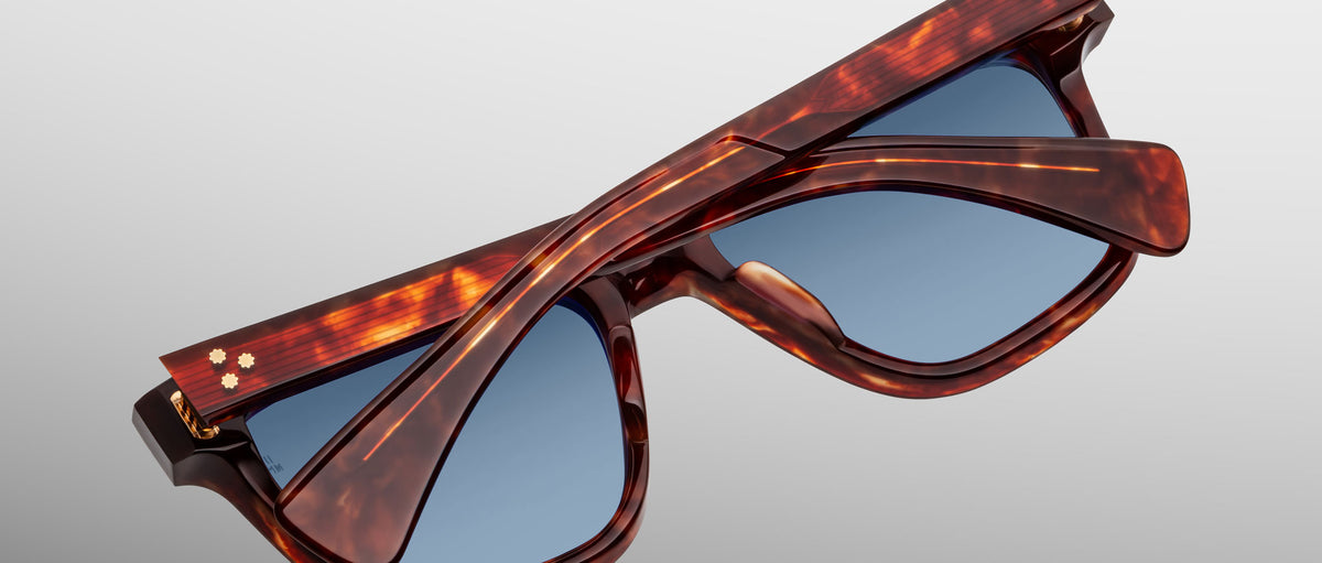 A pair of Jacques Marie Mage Lankaster tortoiseshell sunglasses with blue lenses, folded flat with one temple arm resting atop the other, set against a light gray background.