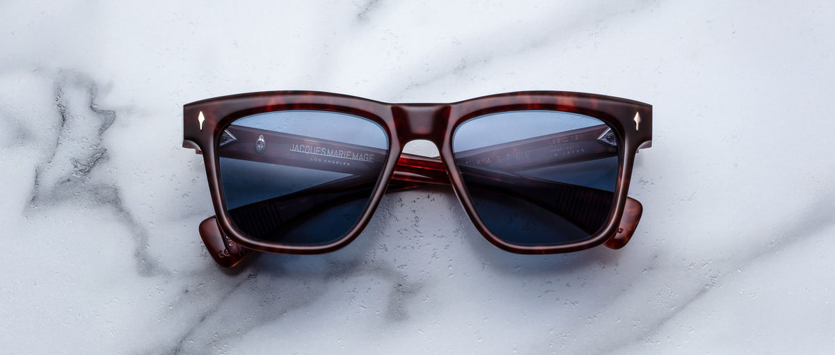 A pair of Jacques Marie Mage Lankaster rectangular tortoiseshell sunglasses with dark lenses rests on a white marble surface.