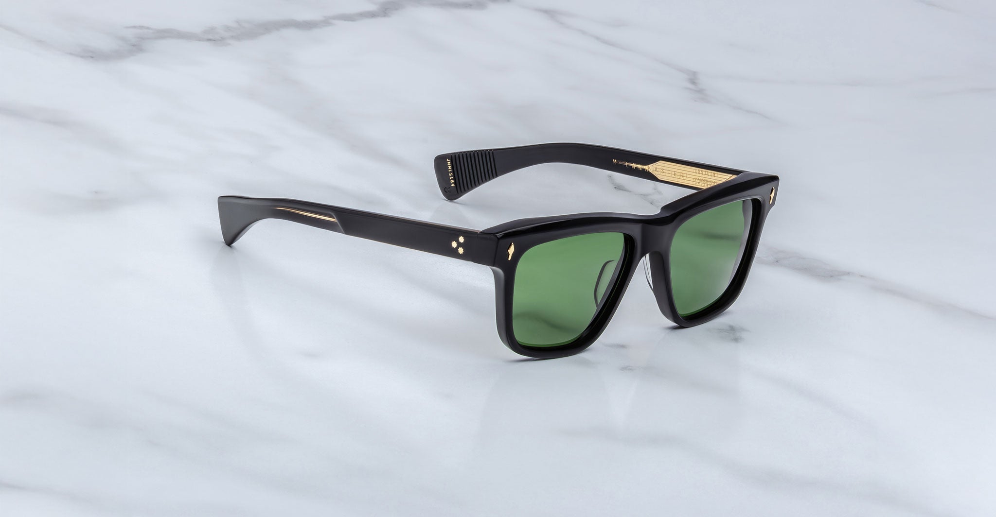 A pair of Jacques Marie Mage Lankaster black sunglasses with green lenses and gold temple accents, displayed on a white marble surface.