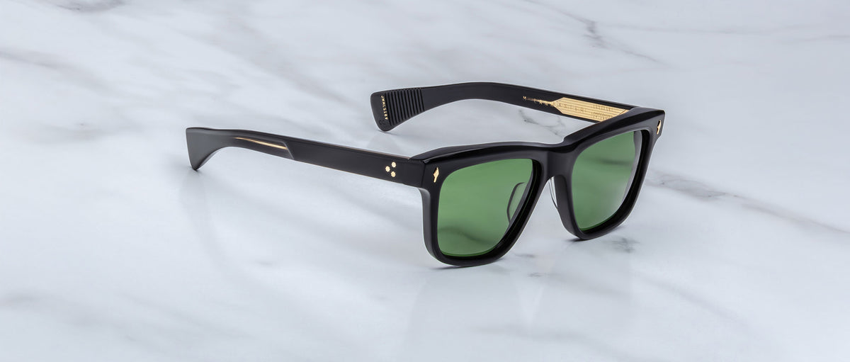 A pair of Jacques Marie Mage Lankaster black sunglasses with green lenses and gold temple accents, displayed on a white marble surface.