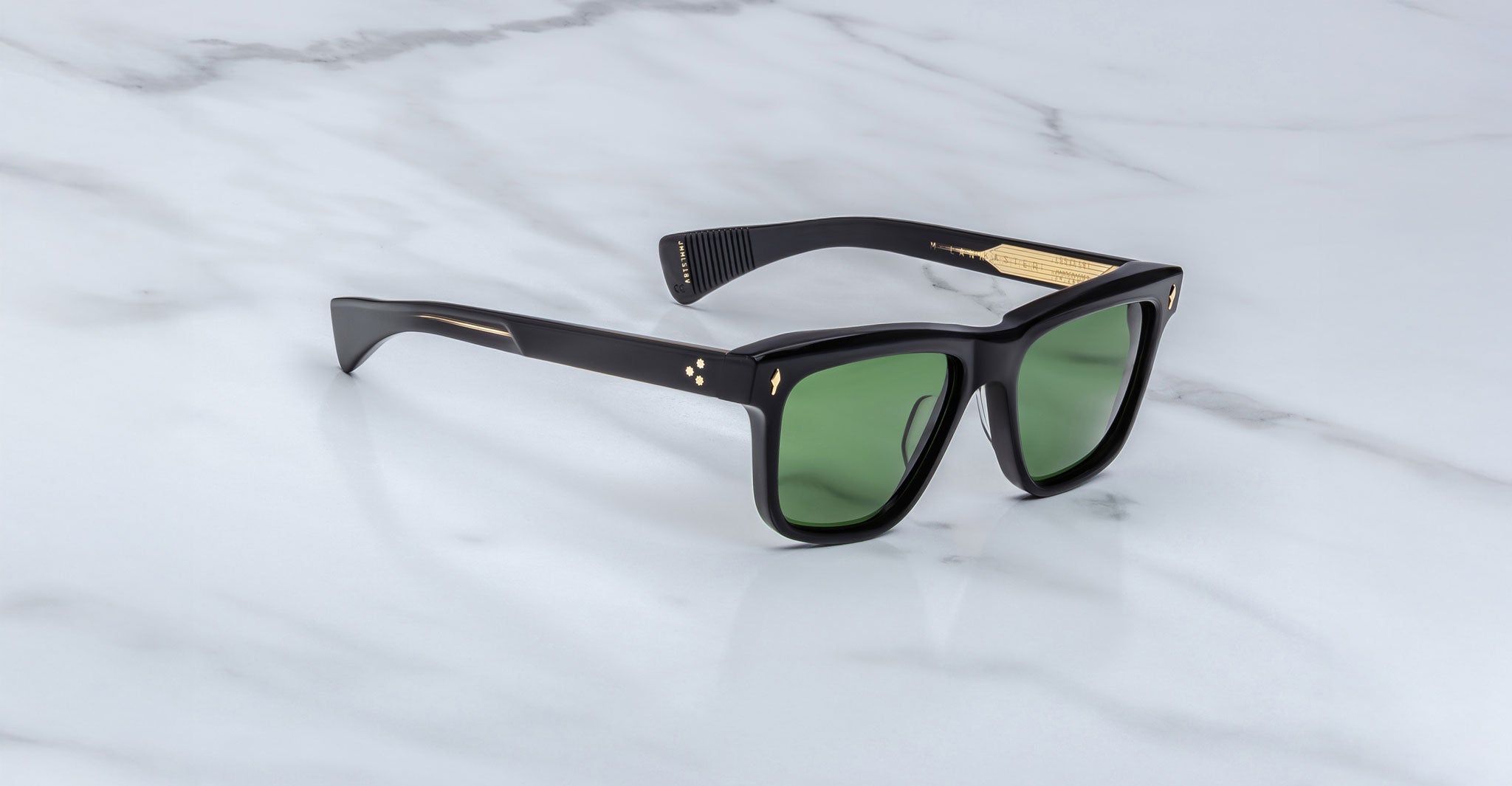 A pair of Jacques Marie Mage Lankaster black sunglasses with green lenses and gold temple accents, displayed on a white marble surface.