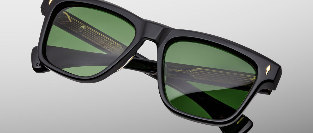 The Jacques Marie Mage Lankaster sunglasses feature a black rectangular frame with green-tinted lenses, gold accents at the front corners, and visible gold detailing on the inner arms, displayed on a light gray background.