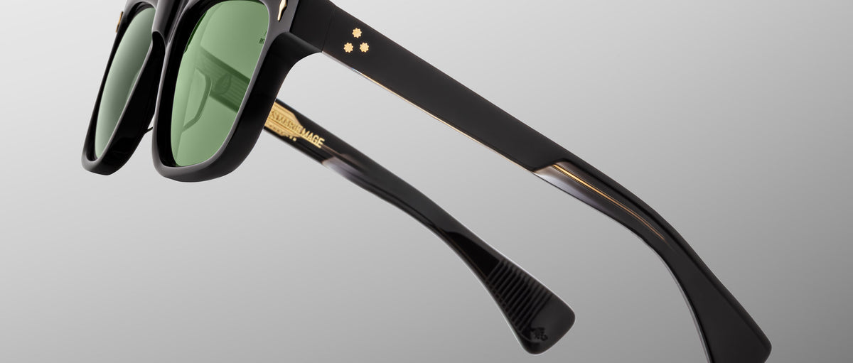 The Jacques Marie Mage Lankaster sunglasses feature black frames with green lenses, gold star-shaped accents, and thick angular arms, displayed against a plain light gray background.