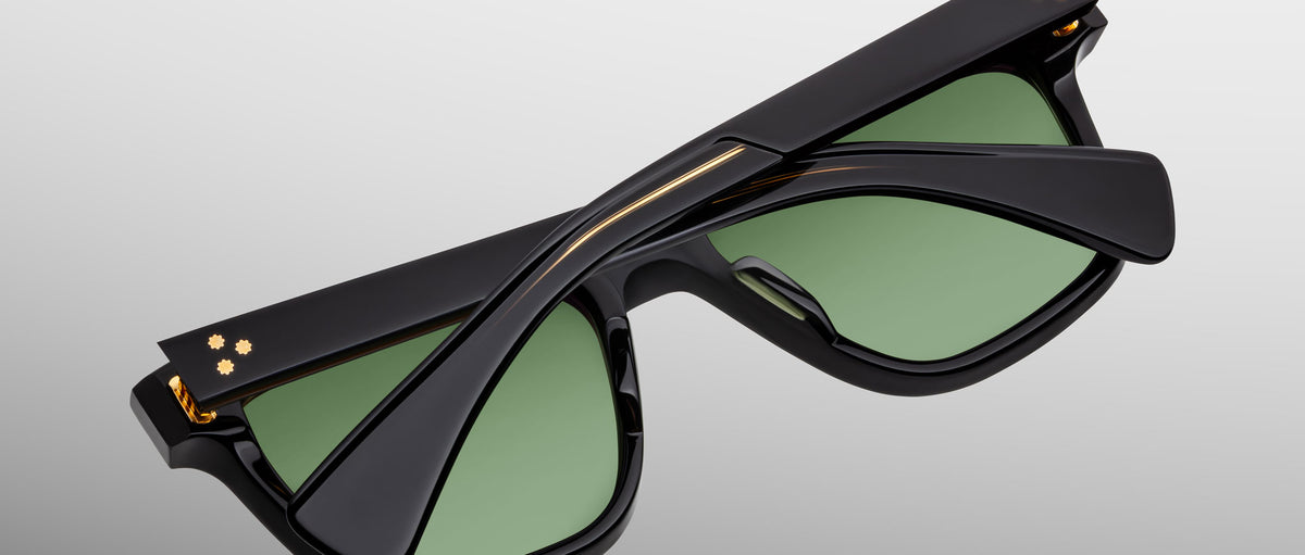 The Jacques Marie Mage Lankaster sunglasses are shown folded neatly, featuring black frames with green lenses and gold accents near the hinges, set against a plain light gray background.