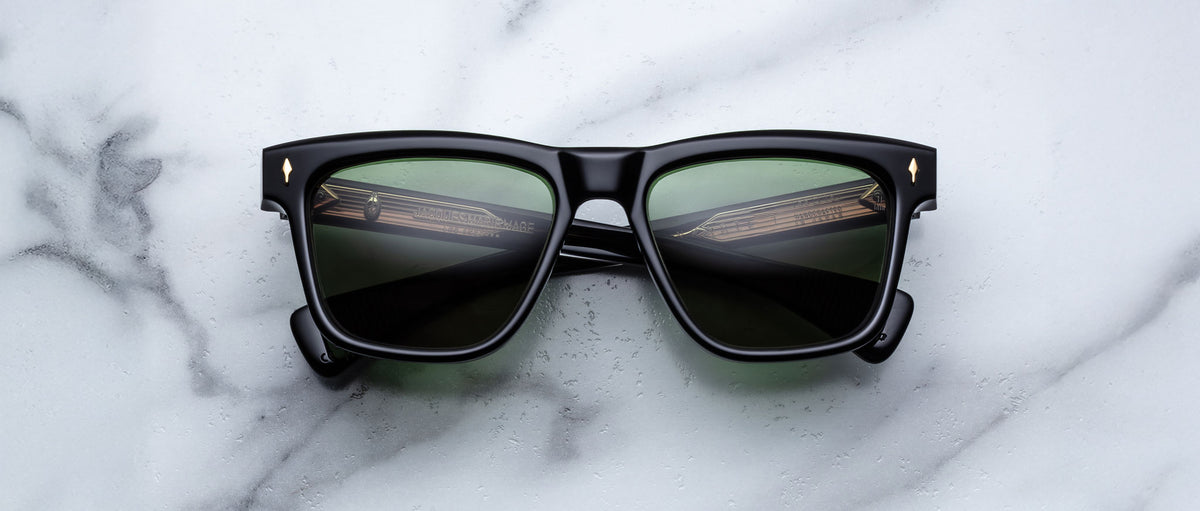 A pair of Jacques Marie Mage Lankaster black rectangular sunglasses with dark green lenses rests on a white marble surface.