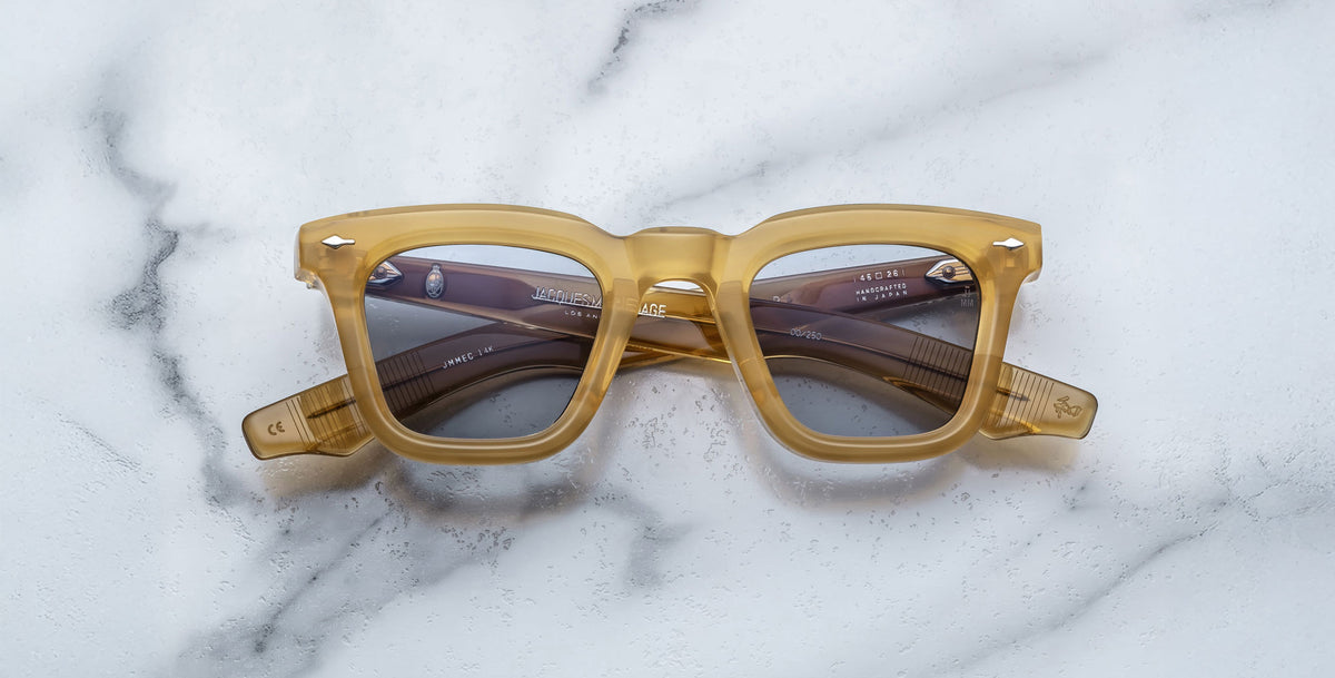 Jacques Marie Mage Leclair eyeglasses with yellow translucent thick frames and gray tinted lenses rest on a white marble surface, viewed from above.