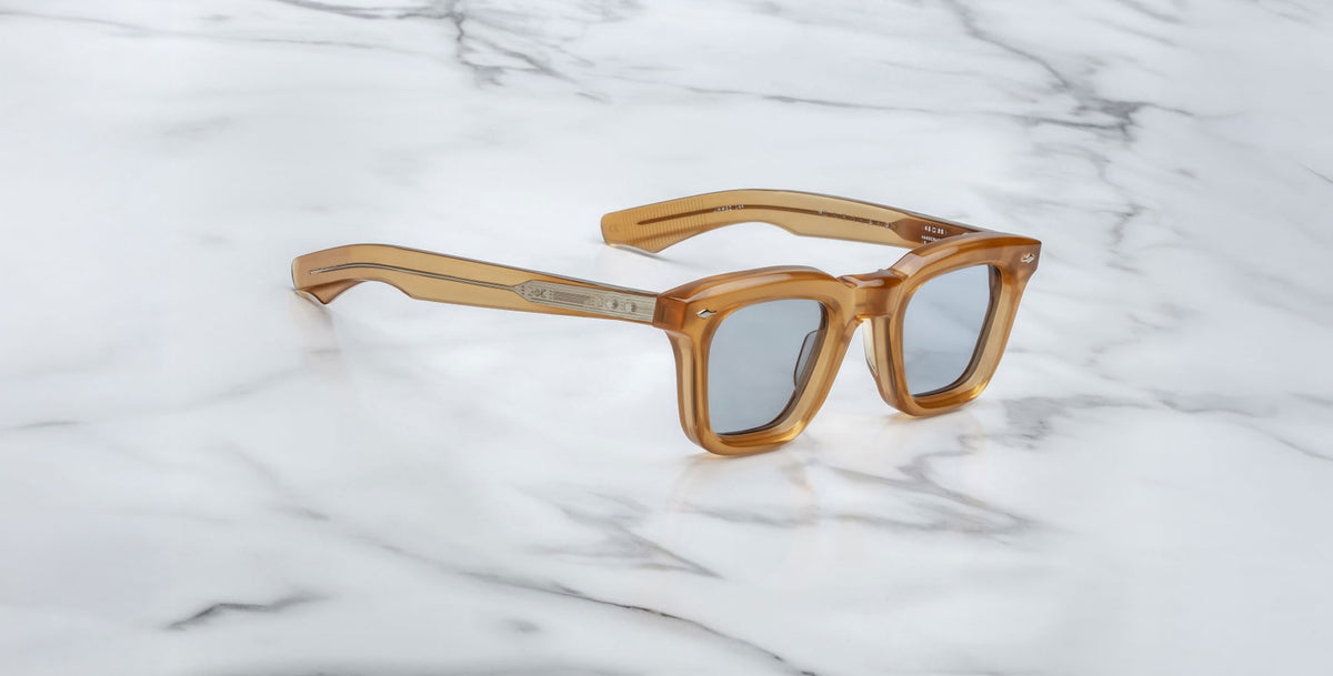 The Jacques Marie Mage Leclair sunglasses, featuring square thick translucent amber frames with silver accents, are displayed on a white marble surface.