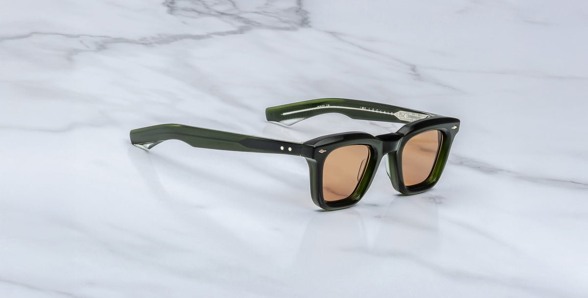 A pair of Jacques Marie Mage Leclair sunglasses with rectangular, dark green frames, brown-tinted lenses, and silver accents by the hinges rests on a white marble surface.