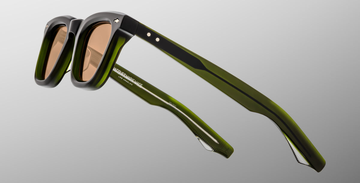 The Jacques Marie Mage Leclair sunglasses, featuring black and green frames with brown tinted lenses, are shown close up on a light grey background to highlight their distinctive frame and arm details.