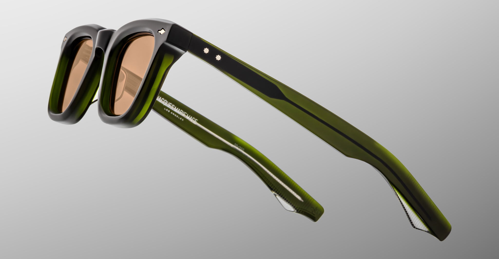 The Jacques Marie Mage Leclair sunglasses, featuring black and green frames with brown tinted lenses, are shown close up on a light grey background to highlight their distinctive frame and arm details.
