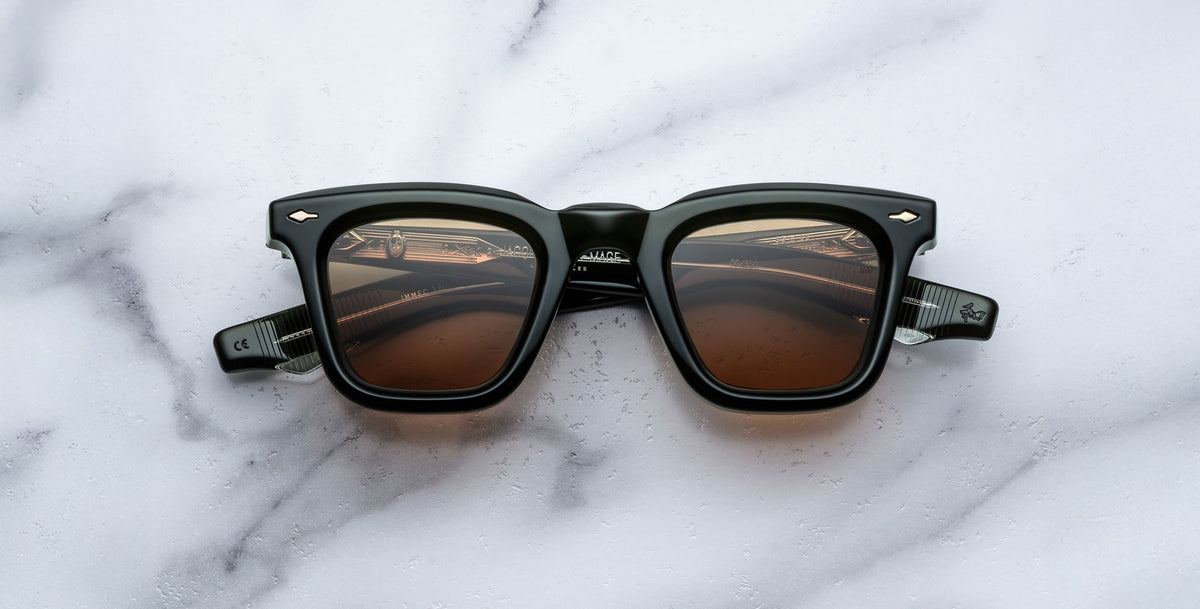 A pair of Jacques Marie Mage Leclair sunglasses with black frames and brown tinted lenses is placed on a white marble surface, viewed from above.