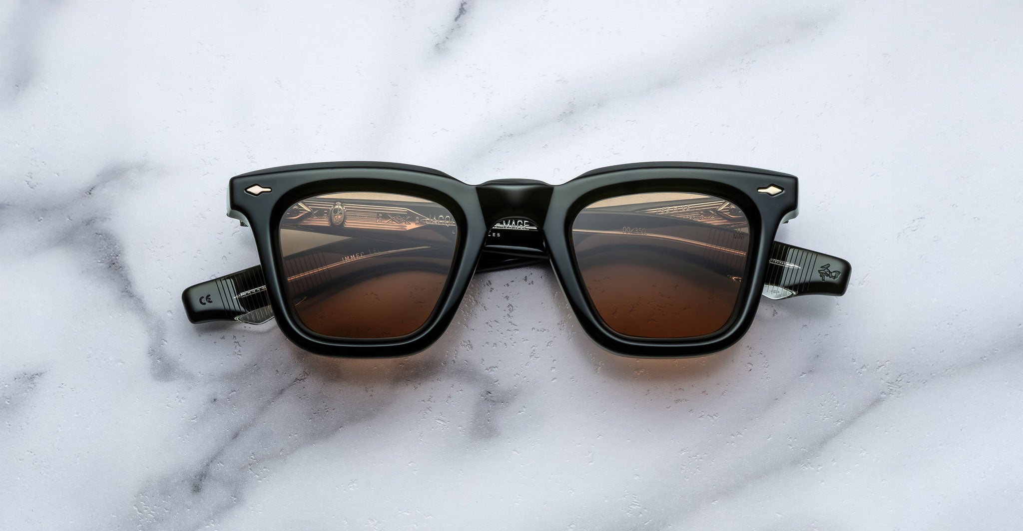 A pair of Jacques Marie Mage Leclair sunglasses with black frames and brown tinted lenses is placed on a white marble surface, viewed from above.