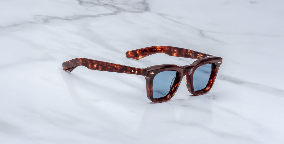 The Jacques Marie Mage Leclair sunglasses, featuring square tortoiseshell frames and blue-tinted lenses, rest on a white marble surface.