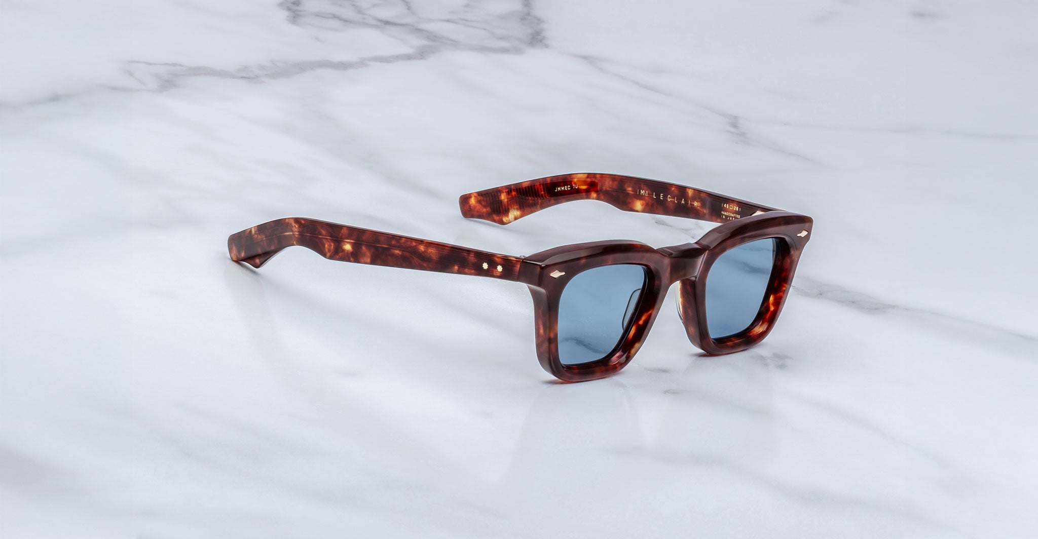 The Jacques Marie Mage Leclair sunglasses, featuring square tortoiseshell frames and blue-tinted lenses, rest on a white marble surface.