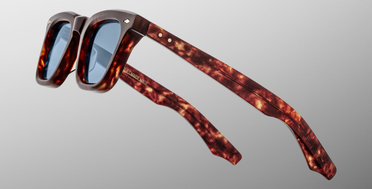 The Jacques Marie Mage Leclair sunglasses feature thick tortoiseshell-patterned frames and blue-tinted lenses, showcased at an angle against a plain light gray background.