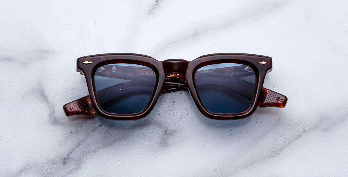 The Jacques Marie Mage Leclair sunglasses, featuring dark brown square frames and blue-tinted lenses, are displayed on a white marble surface.