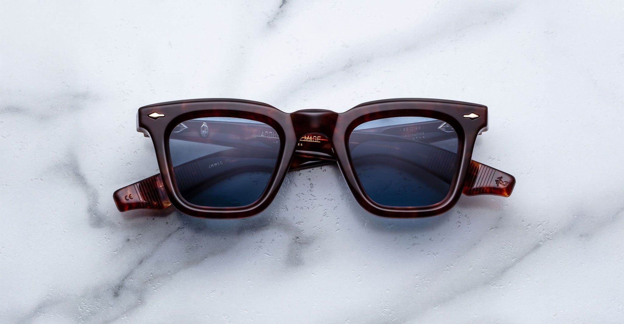 The Jacques Marie Mage Leclair sunglasses, featuring dark brown square frames and blue-tinted lenses, are displayed on a white marble surface.