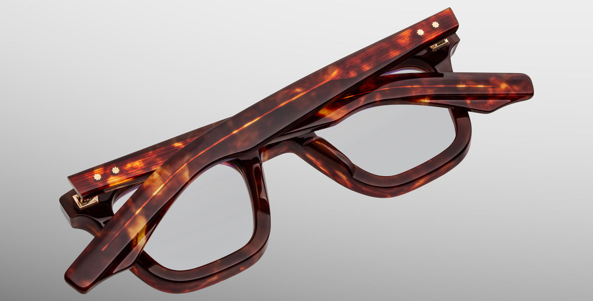 The Jacques Marie Mage Leclair eyeglasses, featuring a tortoiseshell pattern and rectangular lenses, are shown folded on a light gray background.