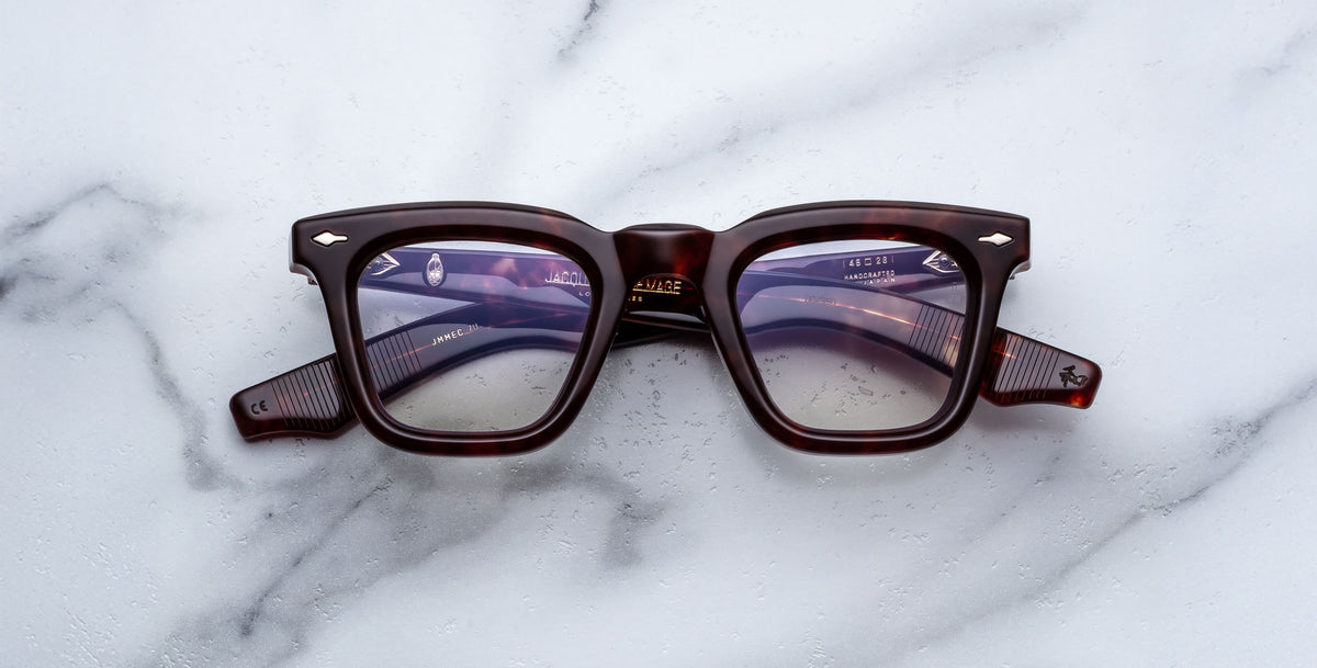 A pair of Jacques Marie Mage Leclair rectangular, thick-framed eyeglasses with a dark tortoiseshell pattern rests on a white marble surface.