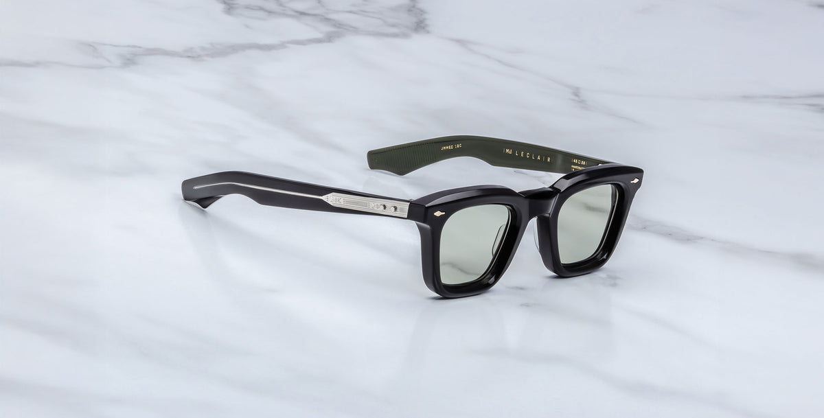 The Jacques Marie Mage Leclair sunglasses feature black rectangular frames with silver temple accents, tinted lenses, and gold text on the inner arms, displayed on a white marble surface.