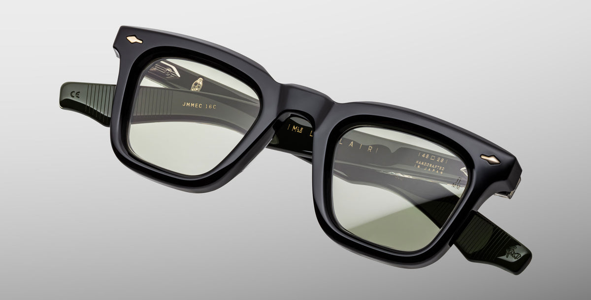 The Jacques Marie Mage Leclair eyeglasses feature thick-rimmed, square black frames with slightly tinted lenses, subtle ridged arms, and gold accents, displayed against a light gray background.