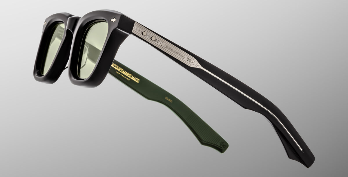 Jacques Marie Mage Leclair sunglasses with black frames, green-tinted lenses, and green inner arms are shown angled on a grey background. The arm has a silver metallic accent and engraved branding.