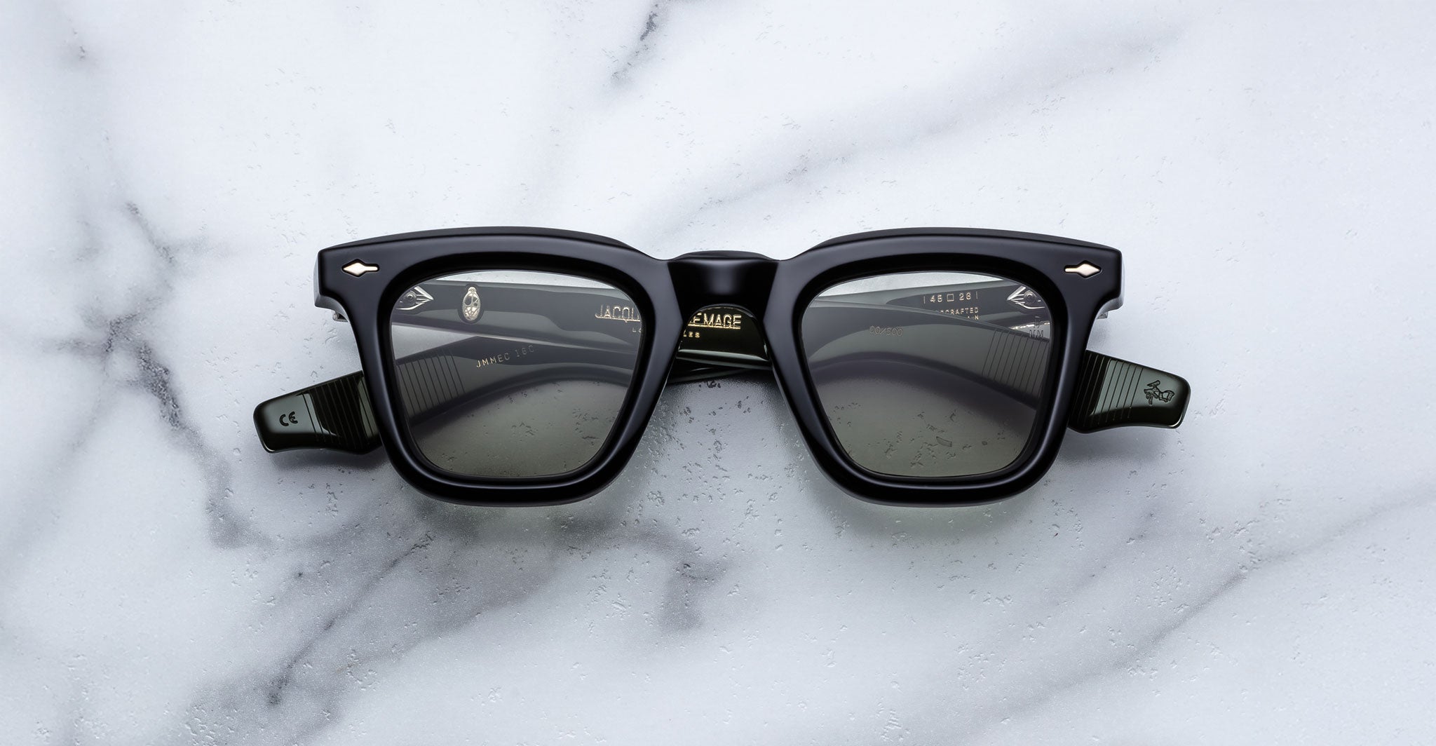 The Jacques Marie Mage Leclair sunglasses, featuring a black rectangular frame with subtle patterned temples and metallic accents near the hinges, rest on a white marble surface. The lenses are dark for a sleek, modern look.