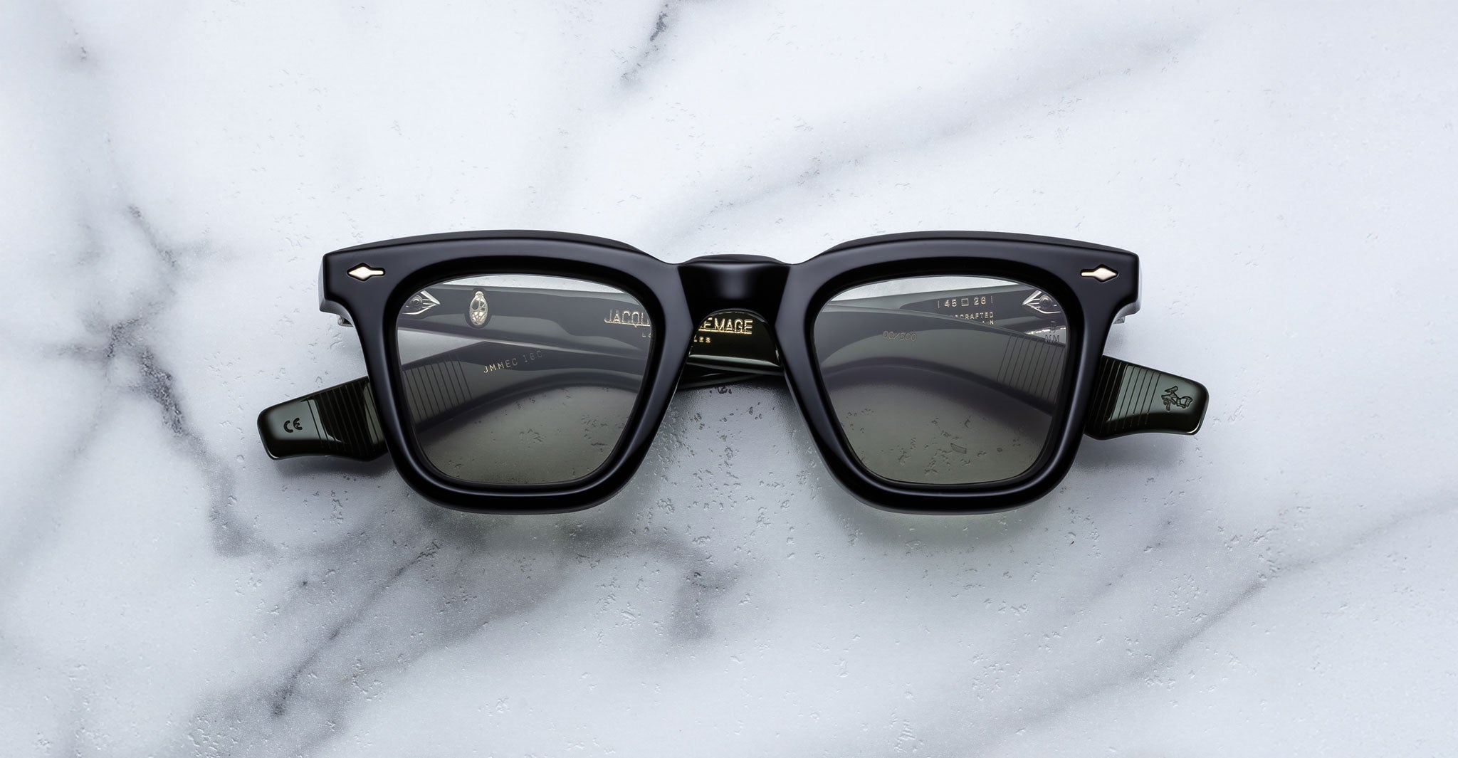 The Jacques Marie Mage Leclair sunglasses, featuring a black rectangular frame with subtle patterned temples and metallic accents near the hinges, rest on a white marble surface. The lenses are dark for a sleek, modern look.