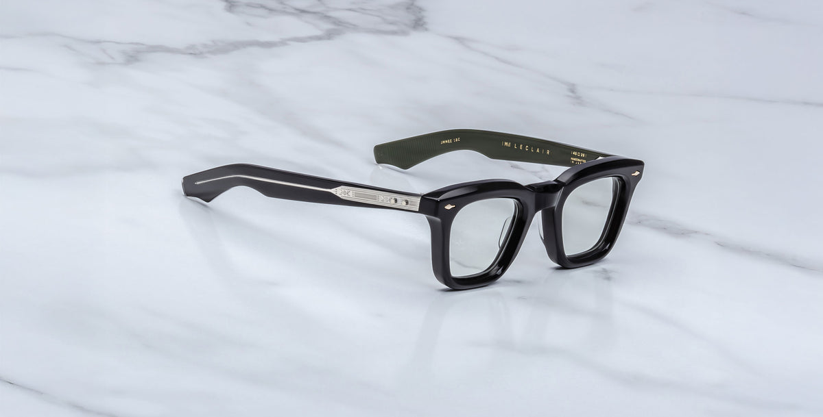 A pair of Jacques Marie Mage Leclair black rectangular eyeglasses with silver accents on the arms rests on a white marble surface. The inner left arm features dark green color and gold text.