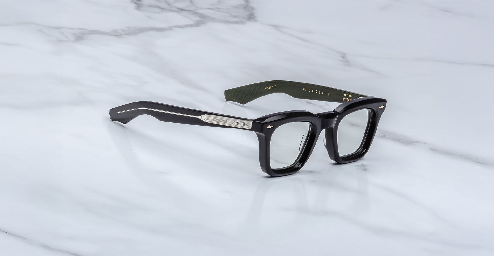 A pair of Jacques Marie Mage Leclair black rectangular eyeglasses with silver accents on the arms rests on a white marble surface. The inner left arm features dark green color and gold text.