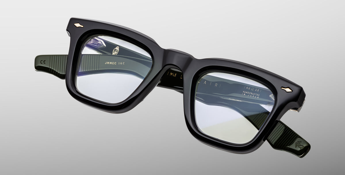 The Jacques Marie Mage Leclair eyeglasses feature black, square frames with clear lenses, showcased at an angle on a light gray background. Gold accents highlight the frame corners and inner arm markings are visible through the lenses.