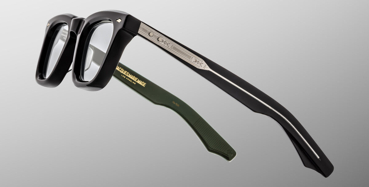 A close-up of Jacques Marie Mage Leclair black eyeglasses with silver metal temples and green inner arms, featuring engraved details and a gold logo inside one arm, set against a gray background.