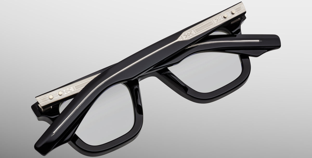 The Jacques Marie Mage Leclair eyeglasses, featuring thick black frames and silver engraved temples, are folded upside down on a light gray surface.