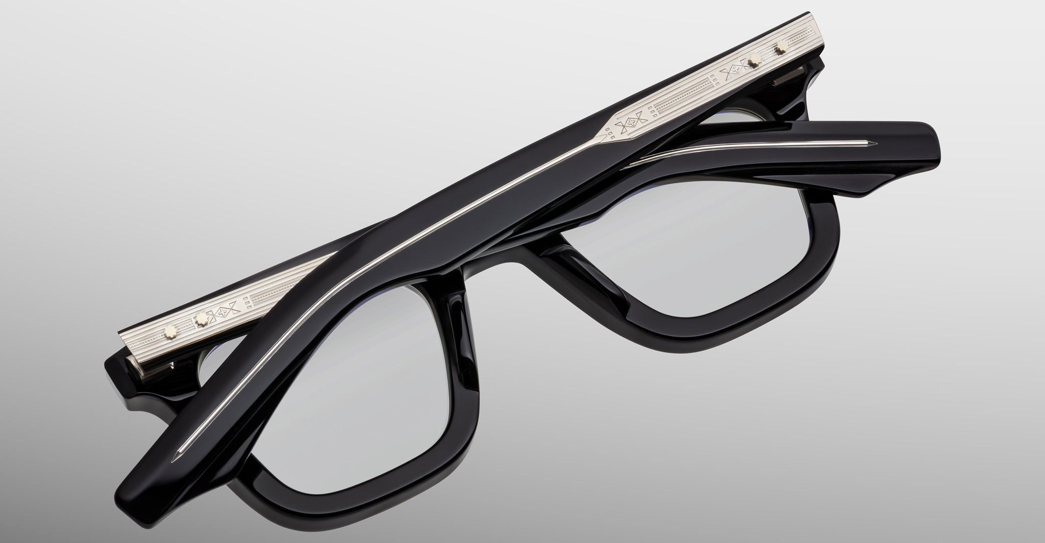 The Jacques Marie Mage Leclair eyeglasses, featuring thick black frames and silver engraved temples, are folded upside down on a light gray surface.