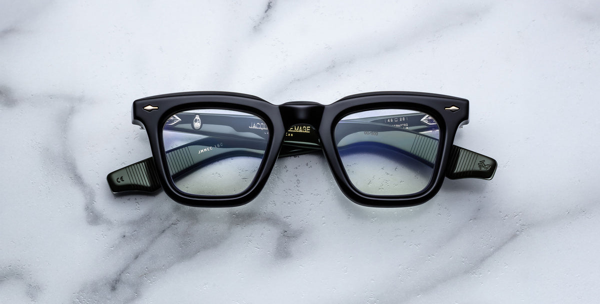 A pair of Jacques Marie Mage Leclair black rectangular eyeglasses with green arms and silver accents rests on a white marble surface, showcasing the brand name and model details inside the frame.