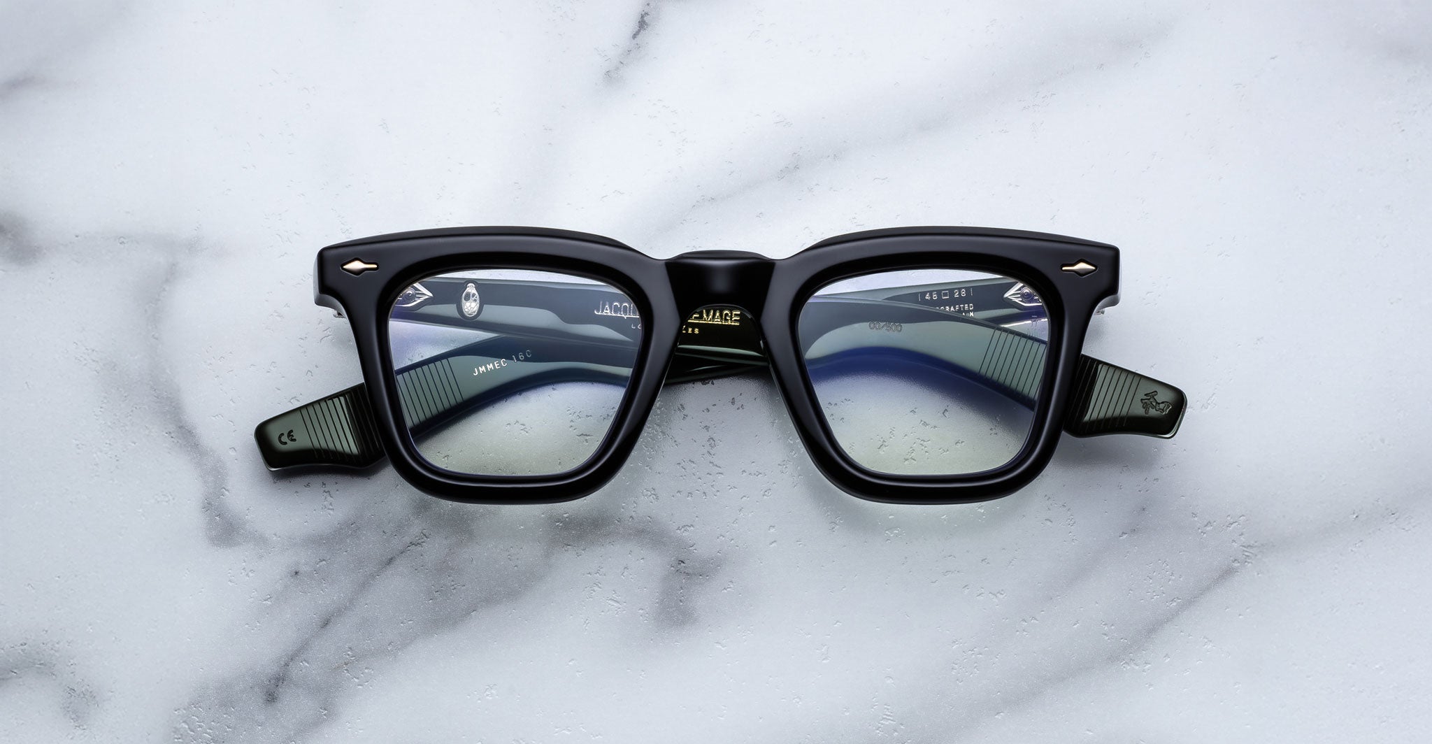 A pair of Jacques Marie Mage Leclair black rectangular eyeglasses with green arms and silver accents rests on a white marble surface, showcasing the brand name and model details inside the frame.