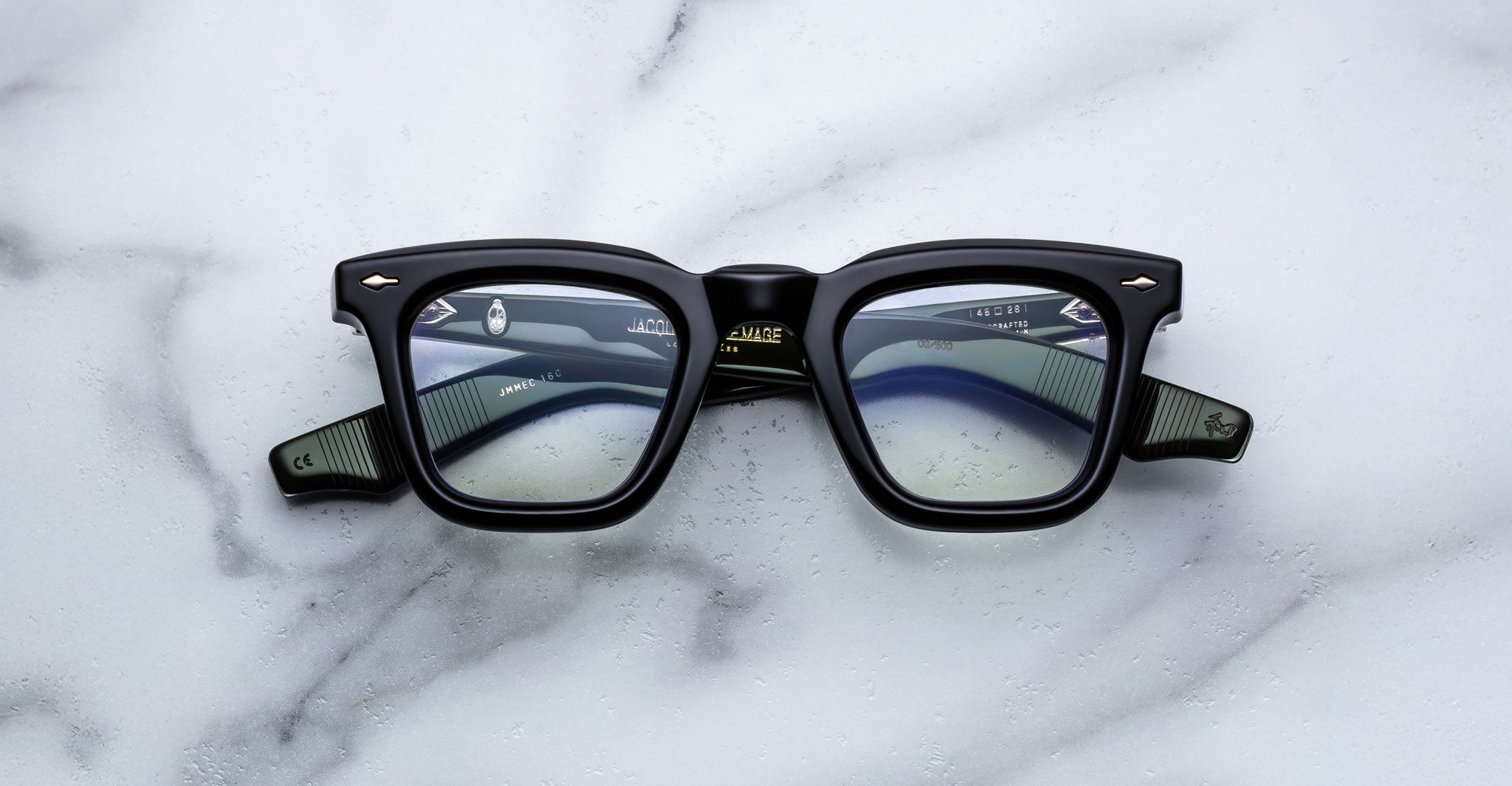 A pair of Jacques Marie Mage Leclair black rectangular eyeglasses with green arms and silver accents rests on a white marble surface, showcasing the brand name and model details inside the frame.